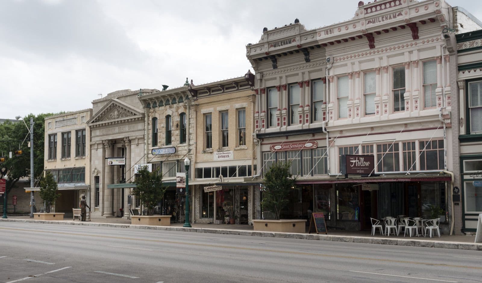 A row of restored 1800s storefronts on the courthouse square in downtown Georgetown, TX, including the ornate cream and red Mileham building dated 1891, with shops, a restaurant patio and young trees along the sidewalk.
